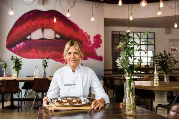 Chef sonriente con paella, perfecta para saber dónde cenar Zaragoza. Mural de labios rojos y luces cálidas crean ambiente.