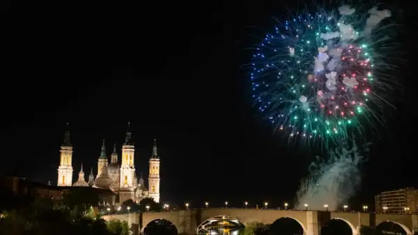 Espectaculares fuegos artificiales iluminan el cielo nocturno sobre el río Ebro, celebrando las fiestas del Pilar. La Basílica del Pilar se alza majestuosa.