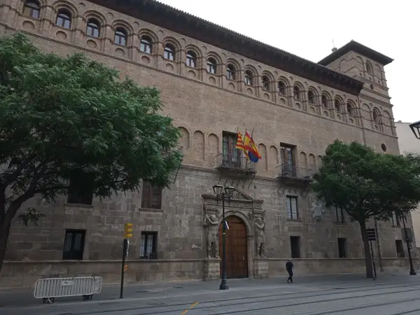 Fachada del palacio de los Luna en Zaragoza, con sus arcos y balcones, flanqueado por árboles verdes.