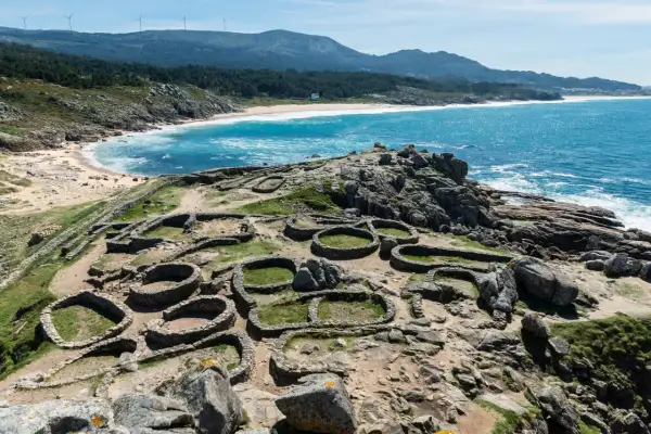 Ruinas circulares de un castro gallego, un lugar histórico que ver en A Coruña, con el mar azul y la costa de fondo.