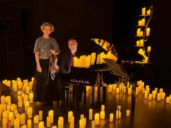 Músicos disfrutan de un concierto con velas. Una violinista y un pianista posan junto a un piano rodeado de velas encendidas.