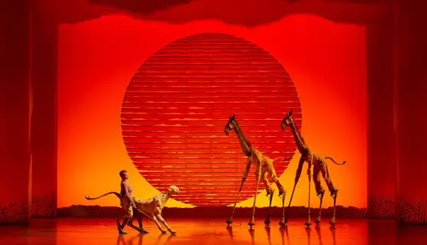 Escena de musicales en Zaragoza: un guepardo y dos jirafas en un escenario con un sol rojo.