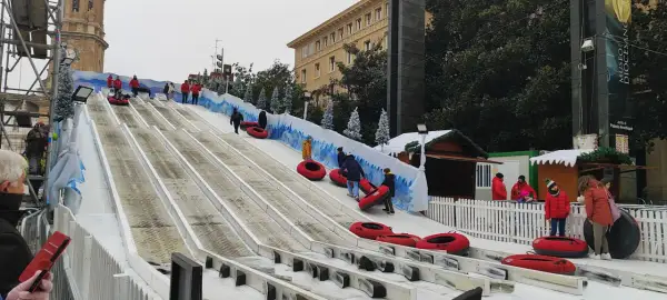 Diversión en los trineos de la Plaza del Pilar. Niños y adultos disfrutan de un tobogán nevado, con la Basílica de Nuestra Señora del Pilar de fondo.