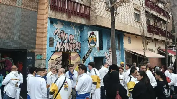 Gente reunida en la calle, algunos con chaquetas blancas y amarillas, celebrando la cincomarzada frente a un edificio con grafitis.