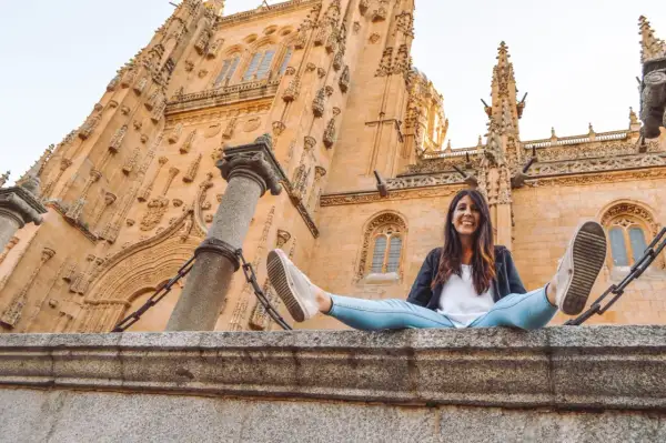 Una mujer sonríe sentada en un muro de piedra frente a la imponente fachada de la Catedral de Salamanca, un lugar clave que ver en Salamanca.