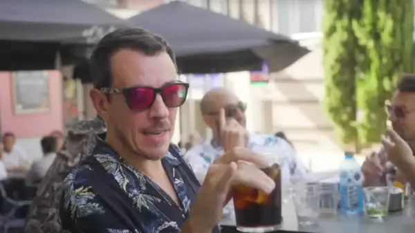 Un hombre con gafas de sol rojas y camisa tropical bebe una bebida en una terraza, quizás disfrutando de un momento de pantomima full Zaragoza.