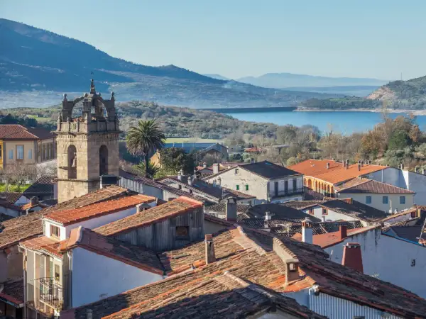 Vista de tejados y un campanario en las Hurdes, con un embalse y montañas al fondo.