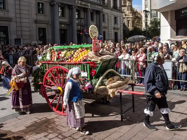 Carro con ofrenda de frutos, pan y verduras, adornado con flores, desfila entre una multitud.