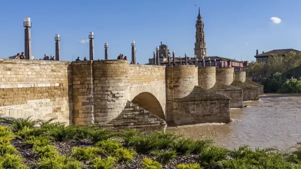El histórico puente de piedra cruza un río caudaloso. En la orilla, gente pasea y se admira la torre de una iglesia.
