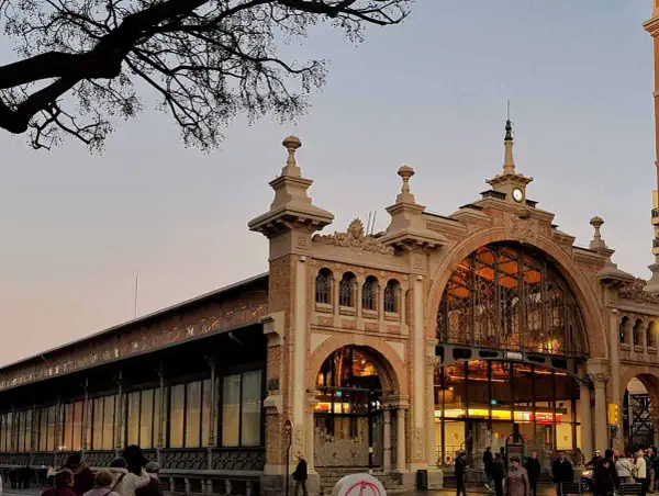 El **mercado central Zaragoza** con su arquitectura de hierro y cristal, iluminado al atardecer. Gente pasea por la entrada principal.