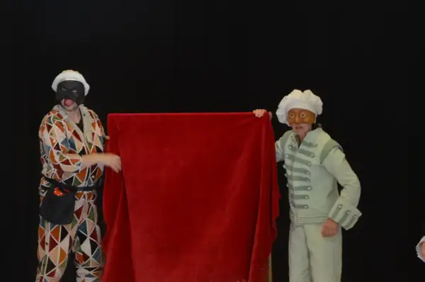 Dos actores con máscaras y trajes de época, uno con un traje de rombos y otro con un traje verde pálido, presentan una tela roja. Parecen parte de una obra de teatro de **els joglars zaragoza**.