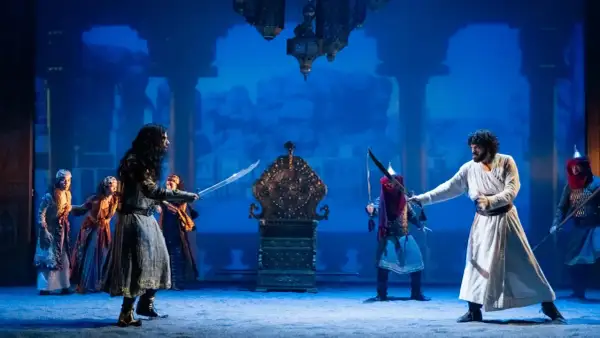 Escena del musical "El Médico" en Zaragoza. Dos hombres se enfrentan con espadas ante un trono dorado, rodeados por otras figuras y un fondo azul.