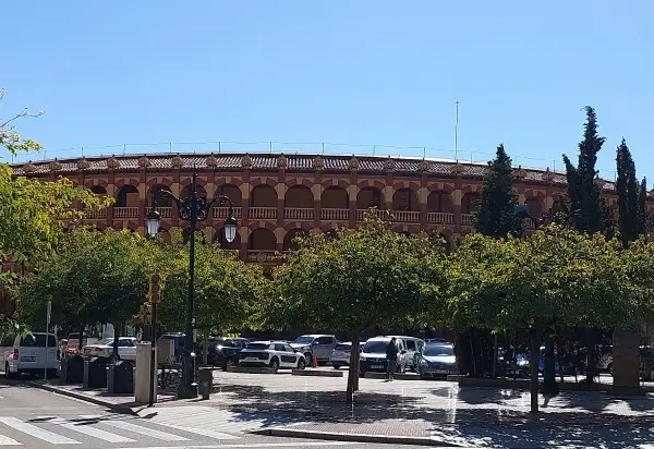 La plaza de toros de Zaragoza, con su arquitectura de arcos y balcones, se alza tras una hilera de árboles frondosos y un aparcamiento con varios coches.