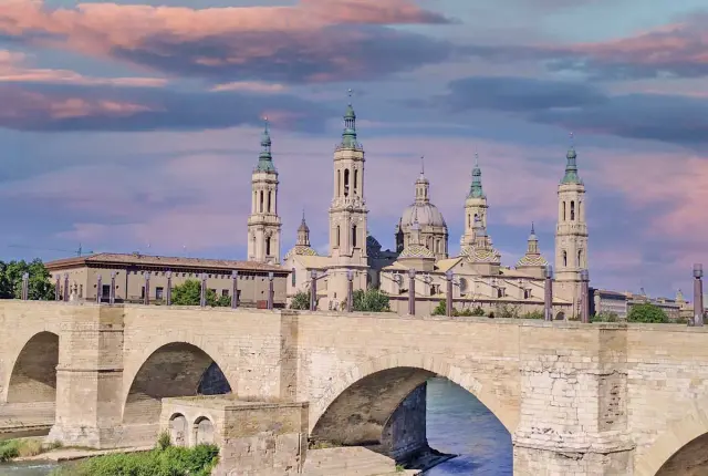 El puente de piedra de Zaragoza, con la Basílica del Pilar al fondo, bajo un cielo rosado.
