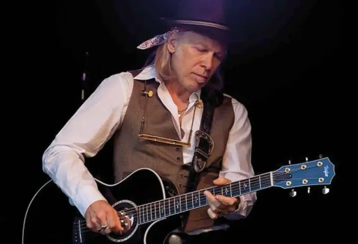 Elliott Murphy Zaragoza, con sombrero y chaleco, toca la guitarra acústica en un concierto.